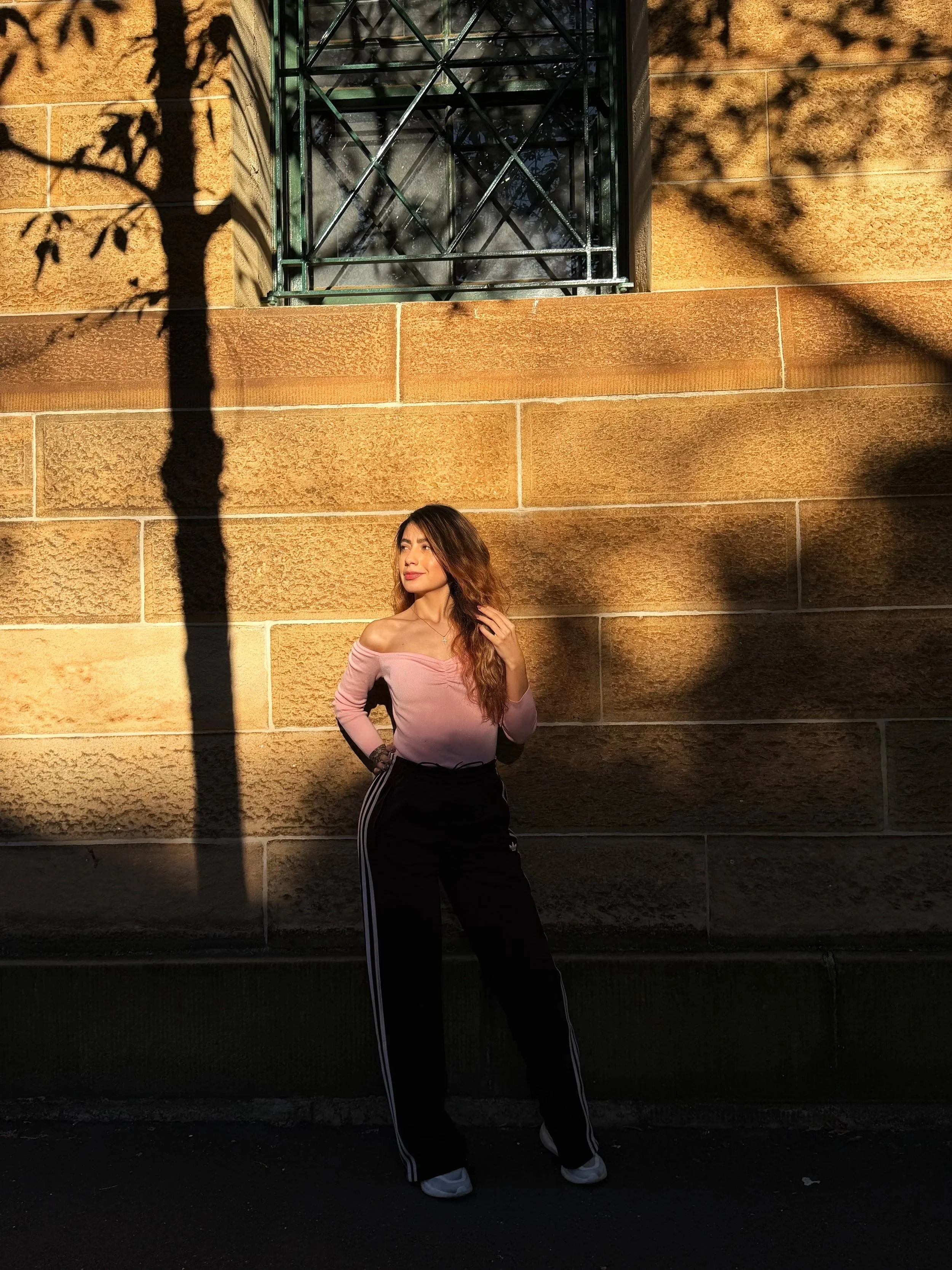Young woman with long hair and a pink off-the-shoulder top standing against a brick wall with shadows of tree branches cast on it, illuminated by sunlight.