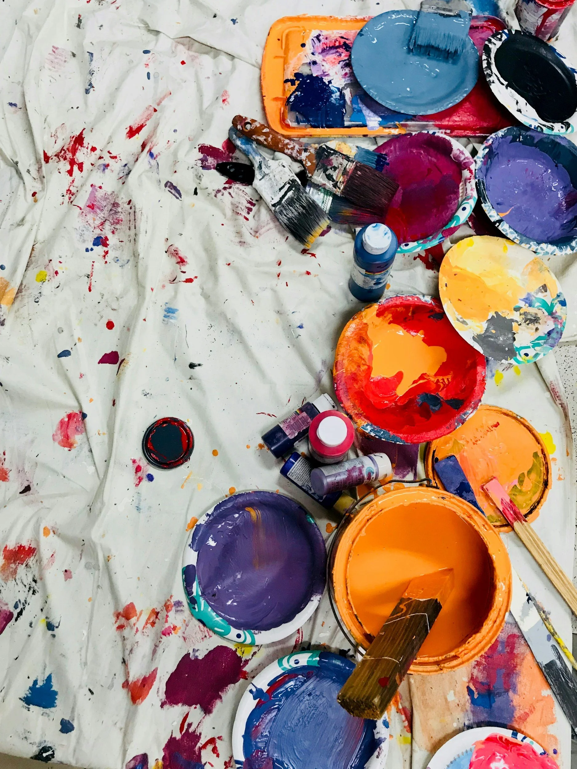 A messy workspace with various paint palettes, brushes, and containers of colorful paint on a covered surface, likely used for painting or art projects.