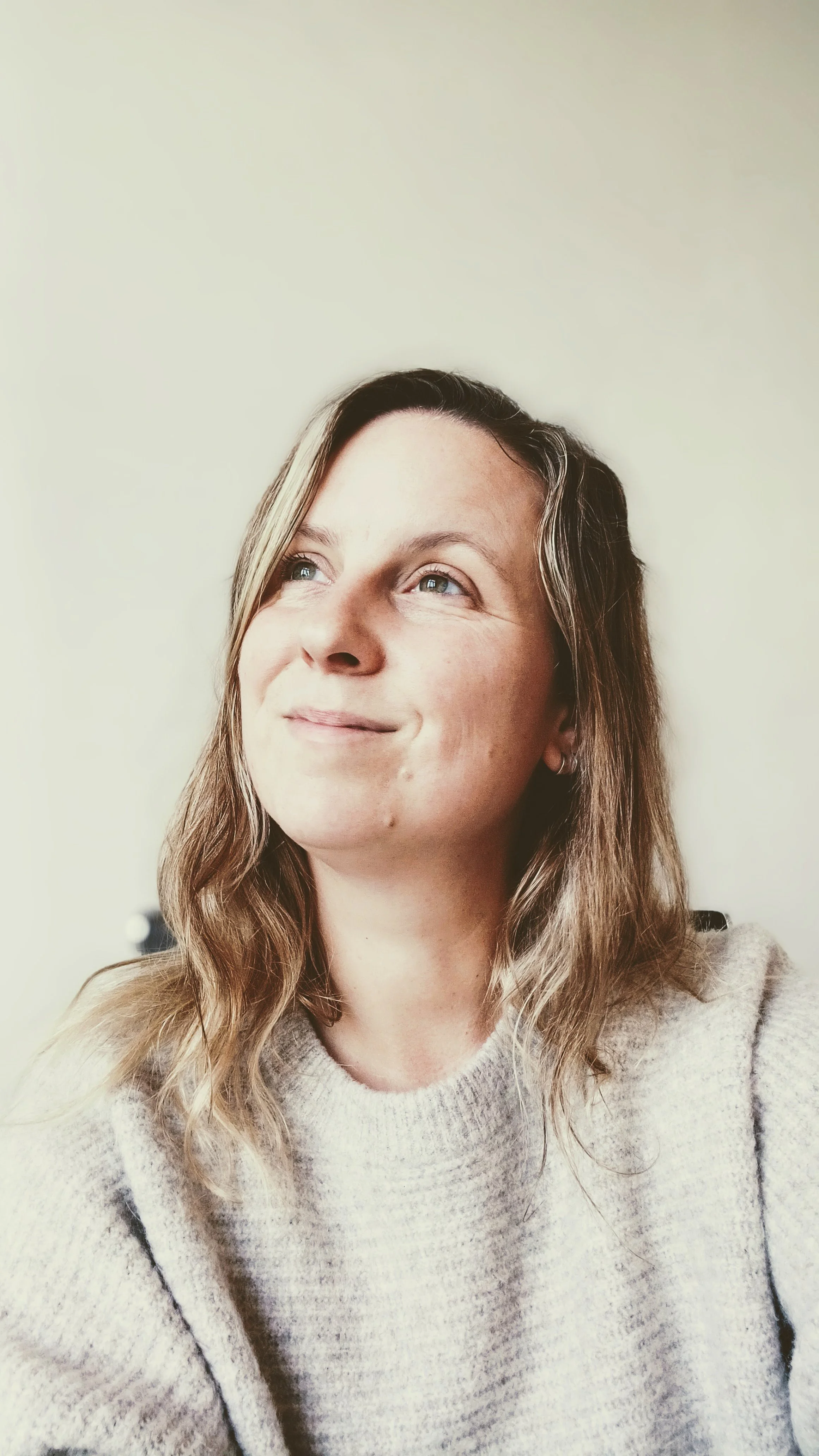 A woman with wavy light brown hair and blue eyes, wearing a beige sweater, looking up and smiling softly, against a plain light-colored wall.