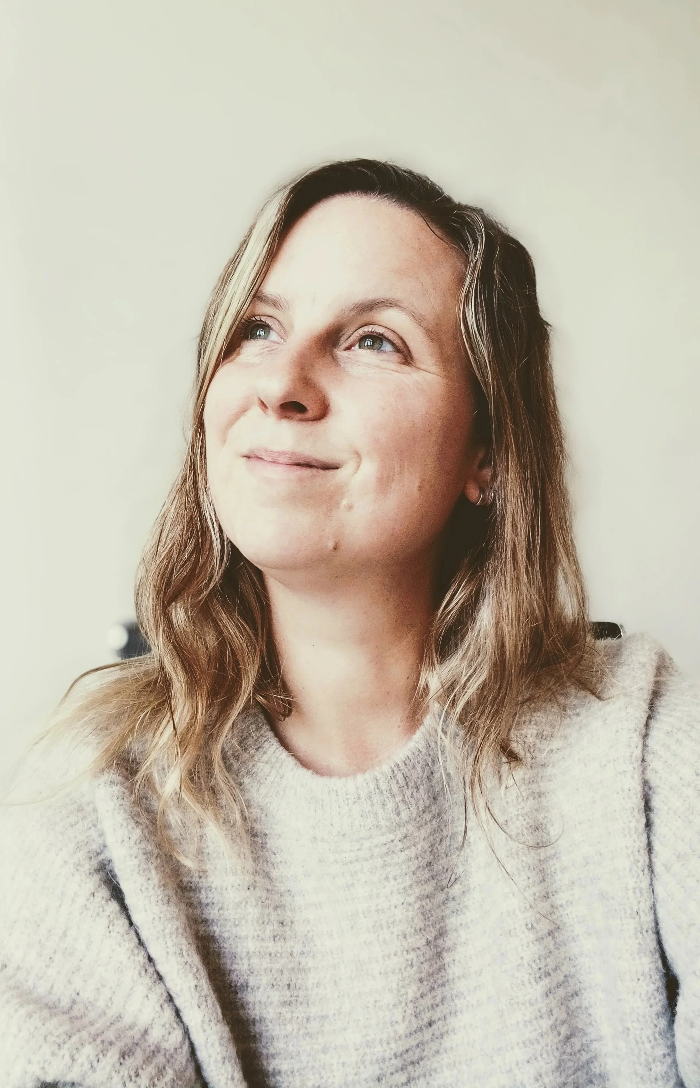 Portret van een vrouw met lange, golvende, lichtbruine haren die naar links kijkt, lachend, in een lichte, gebreide trui, tegen een effen lichte achtergrond.
