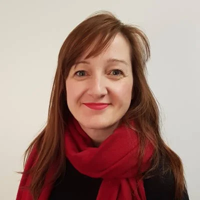 Portrait of a woman with reddish-brown hair, smiling, wearing a red scarf and black top, against a plain white background.