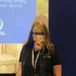 Woman with blonde hair and glasses wearing a black shirt, standing in front of a blue presentation screen.
