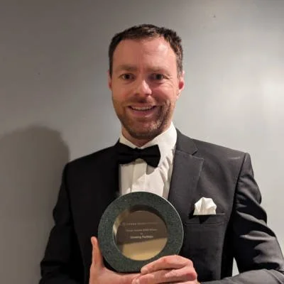 Man in black tuxedo holding an award plaque, smiling, against a plain gray wall.
