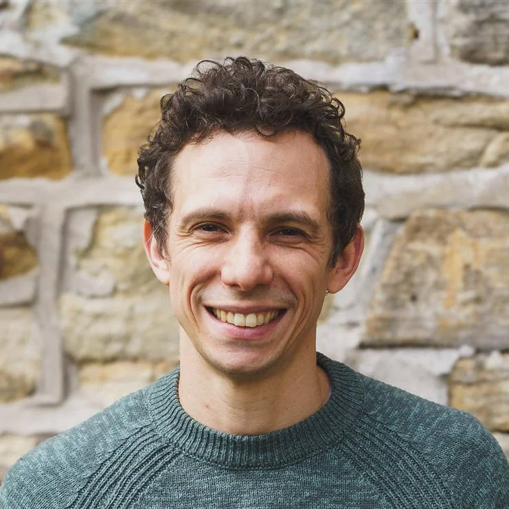A smiling man with curly dark hair standing outdoors in front of a stone wall.