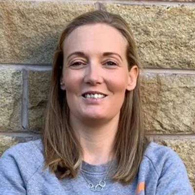 A woman with shoulder-length light brown hair, smiling, standing in front of a beige stone wall, wearing a gray sweatshirt and a silver necklace.