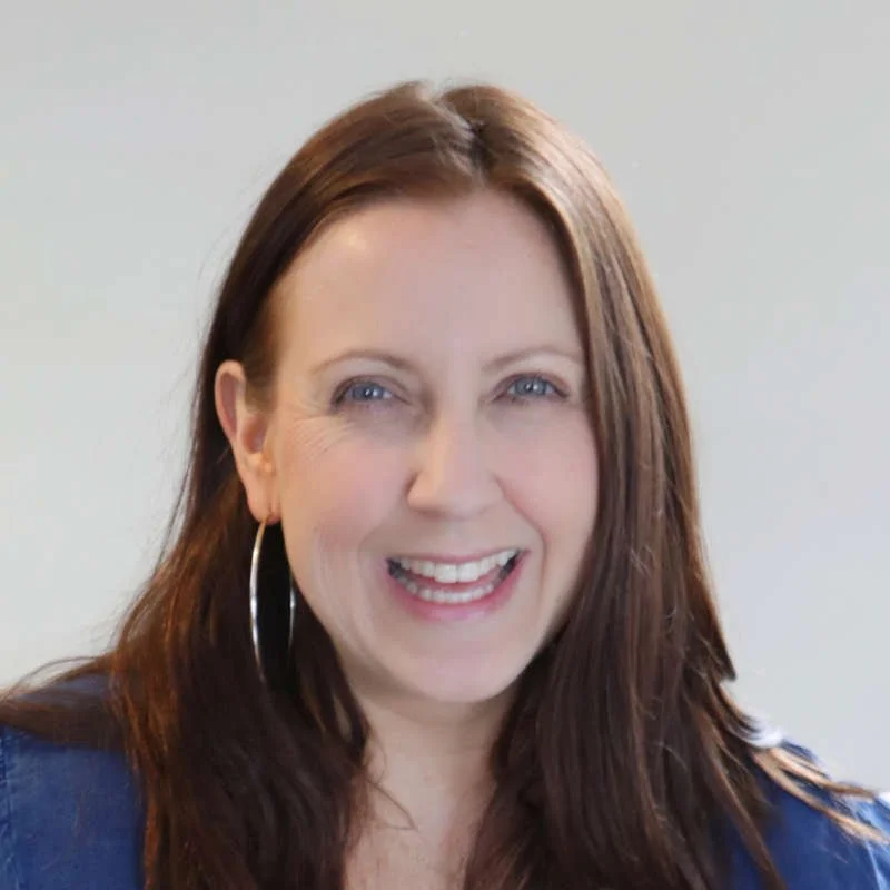 A woman with long, dark brown hair and light blue eyes smiling at the camera, wearing hoop earrings and a dark top, against a plain light background.