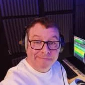 A man with glasses taking a selfie in front of music production equipment, including a computer with colorful display and audio equipment.