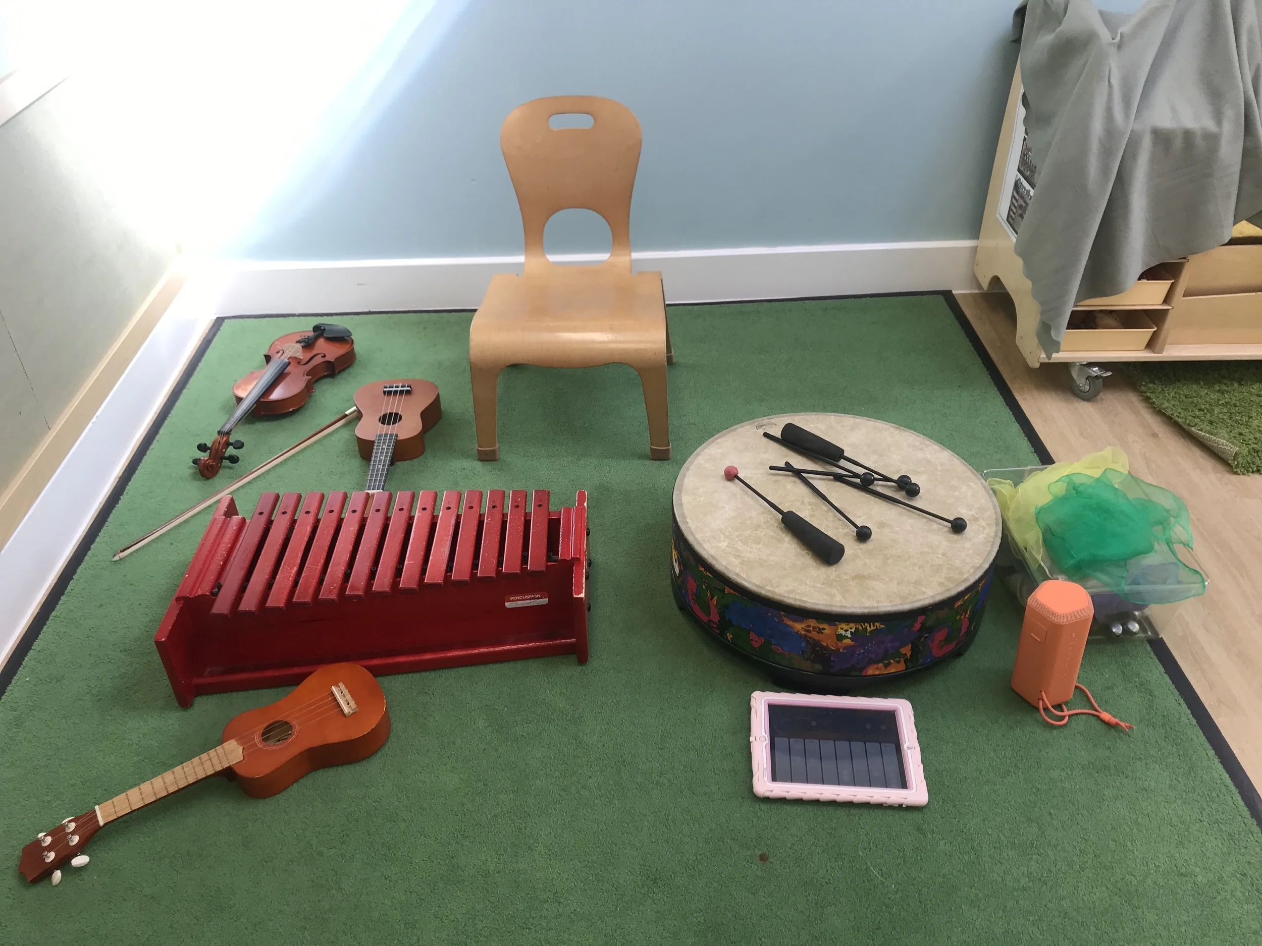 A corner of a children's playroom with musical toys including a small red xylophone, a golden violin and a small guitar, all on green carpet. There is a wooden chair, a drum with drumsticks, a tablet, and a partially visible cupboard with rainbow-colored netting