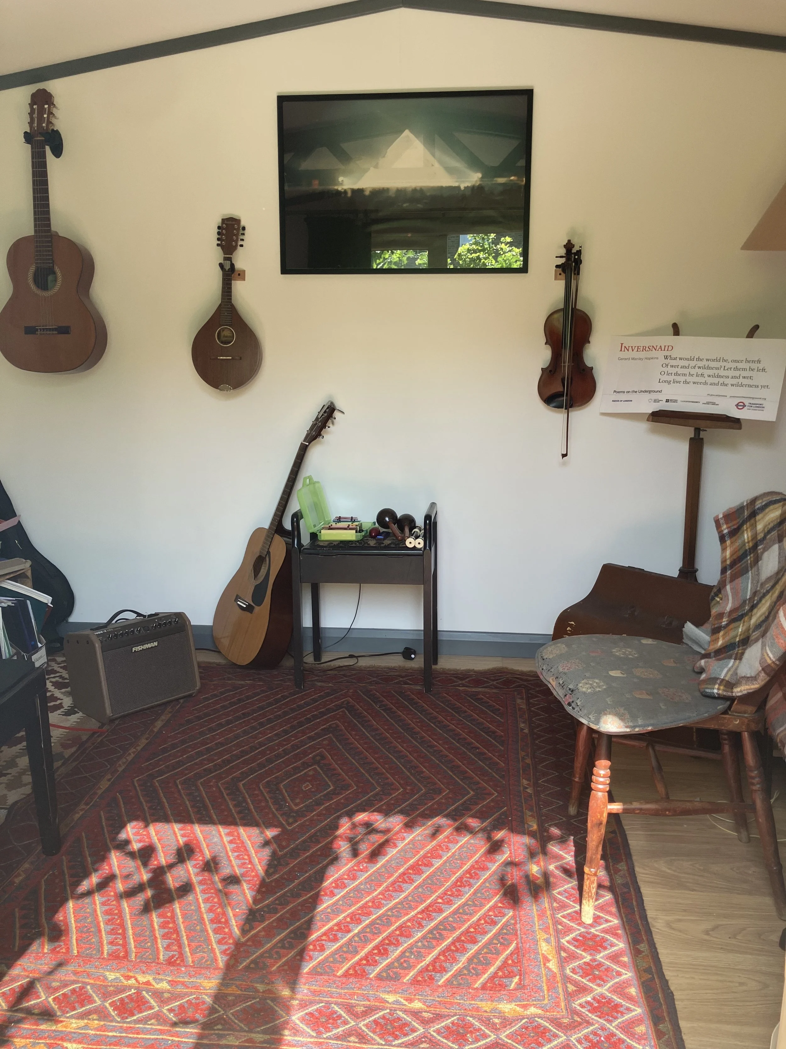 A wall with instruments on hooks including a guitar, a mandolin and a violin. There is a red patterned rug on the floor, a wooden chair with a cushion, and a small table with various musical instruments