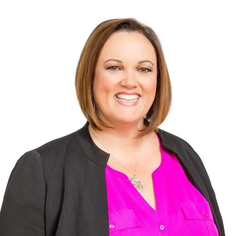 Professional headshot of a smiling woman with a brown bob haircut, wearing a bright pink blouse and dark blazer against a white background