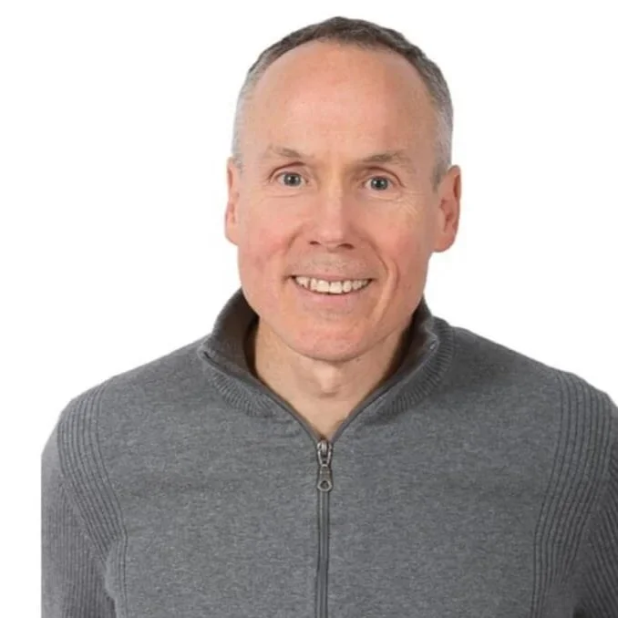 Professional headshot of a smiling man with short grey hair, wearing a grey zip-up sweater against a white background