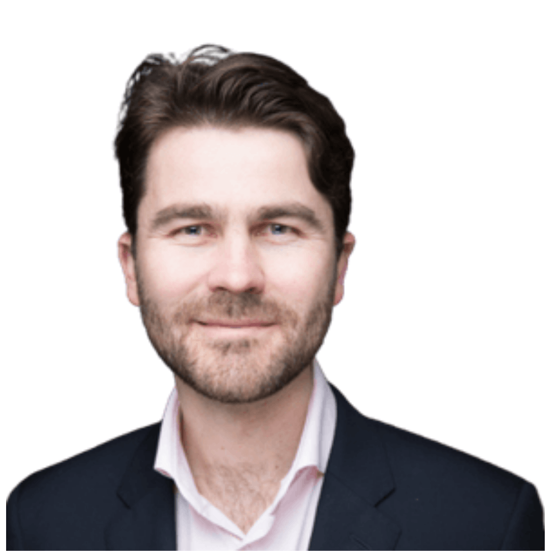 Professional headshot of a smiling man with short dark hair and a beard, wearing a dark blazer over a light pink shirt against a white background