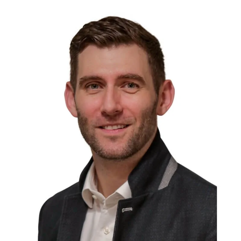 Professional headshot of a smiling man with short brown hair and a trimmed beard, wearing a dark jacket over a white shirt against a white background