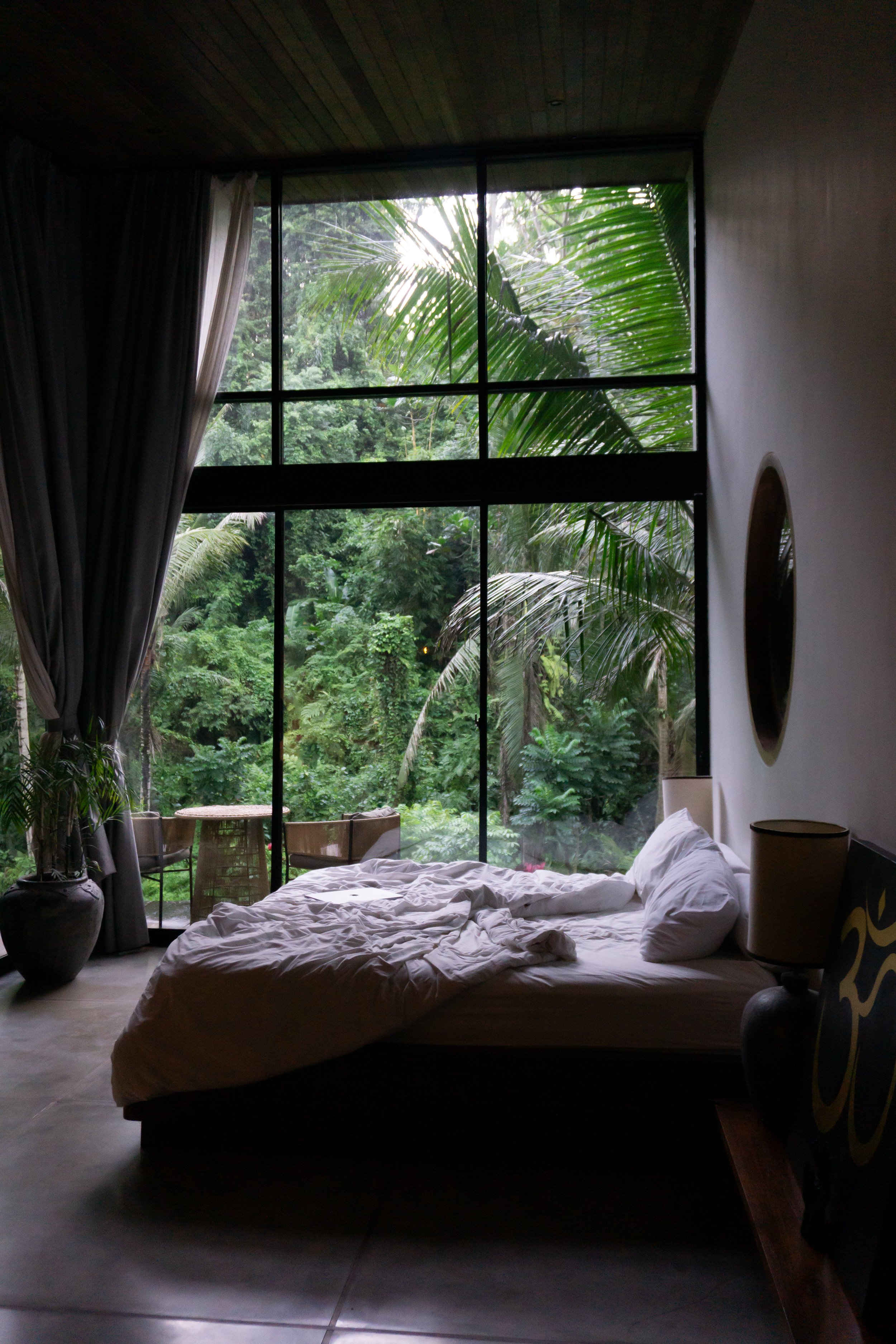 Een slaapkamer met een groot raam dat uitkijkt op een weelderige, groene jungle. Het bed ligt ongeordend met witte lakens en kussens, het lijkt een luxe hotelkamer