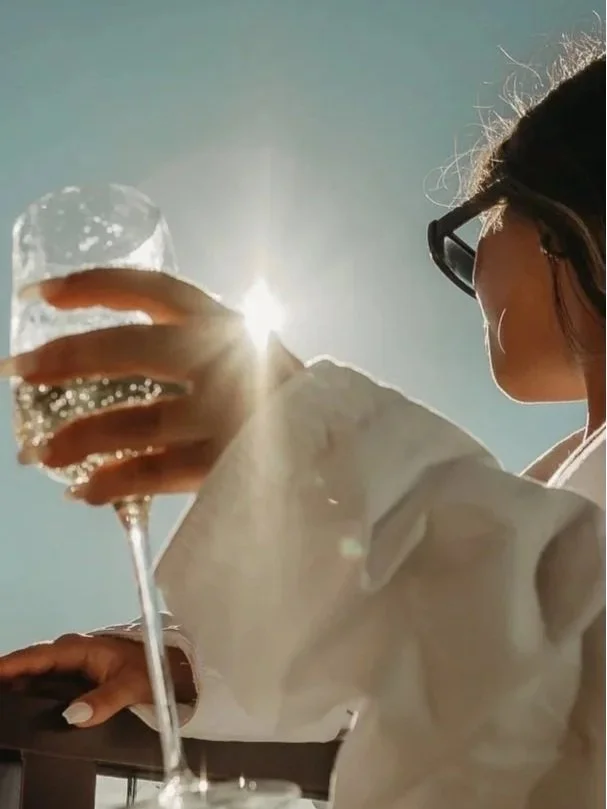 Vrouw met zonnebril houdt een glas champagne omhoog, zonlicht op de achtergrond.
