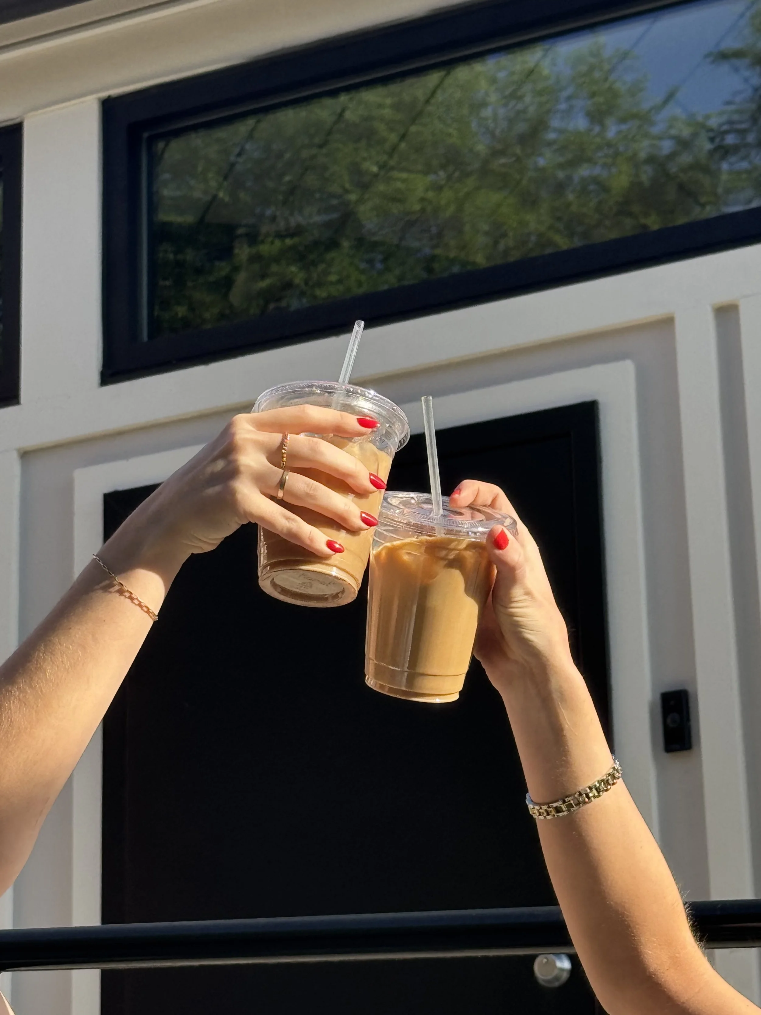 Twee handen met rode nagellak die twee plastic bekers met ijs en drinken omhoog houden, buiten voor een modern gebouw met grote ramen en groene boomreflecties.