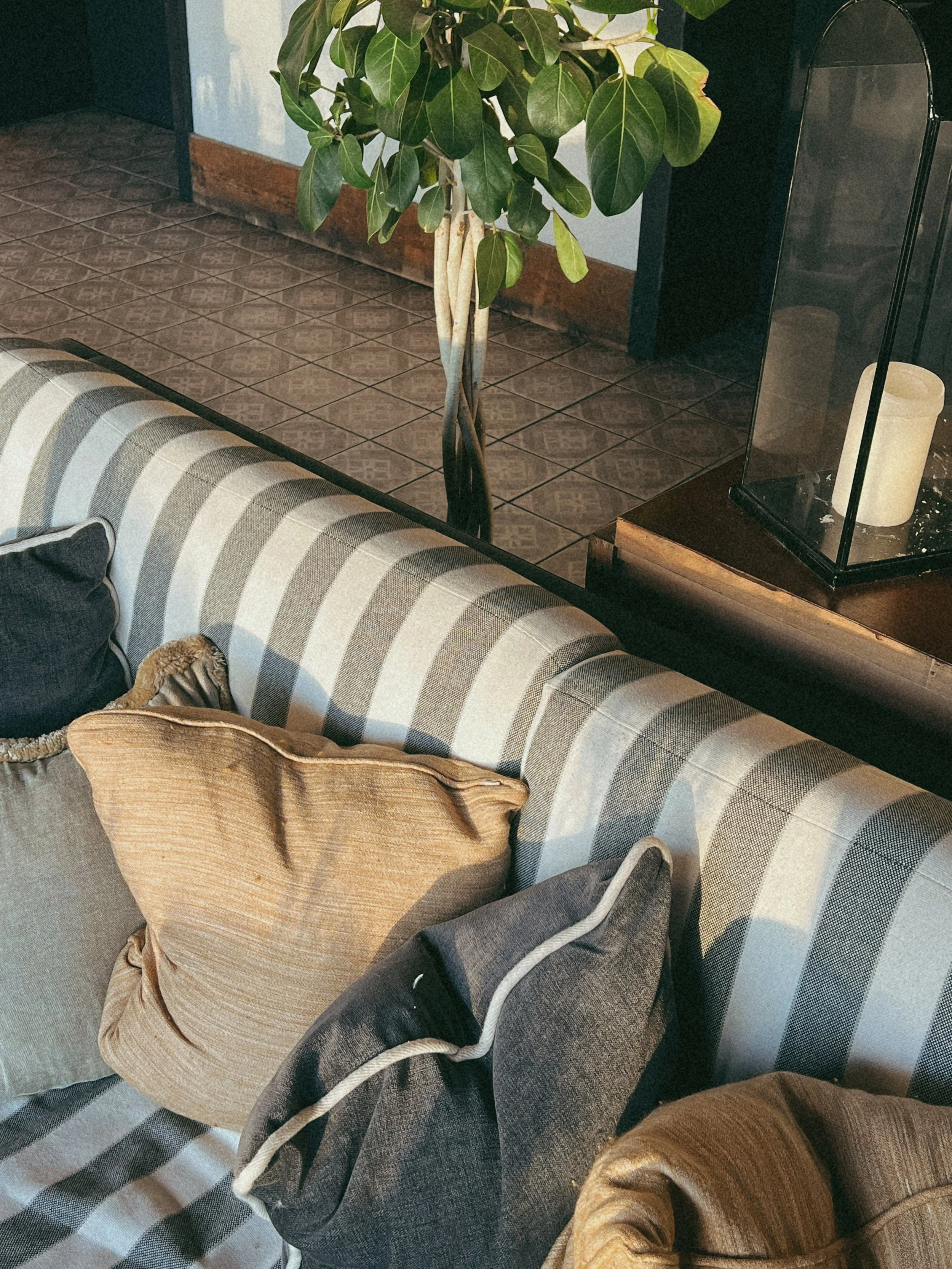 Verschillende stoffen kussens en plaid op een bank met een gestreept patroon, een plant en een glazen lamp op een houten tafel in een woonkamer.
