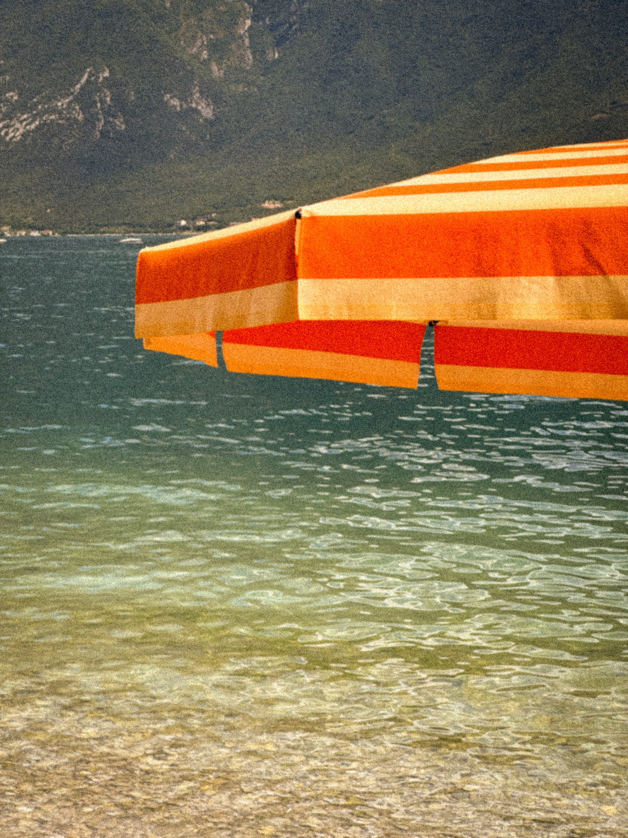 Zeewater met een zandstrand en een uitgevouwen oranje en beige strandparaplu, bergen op de achtergrond.