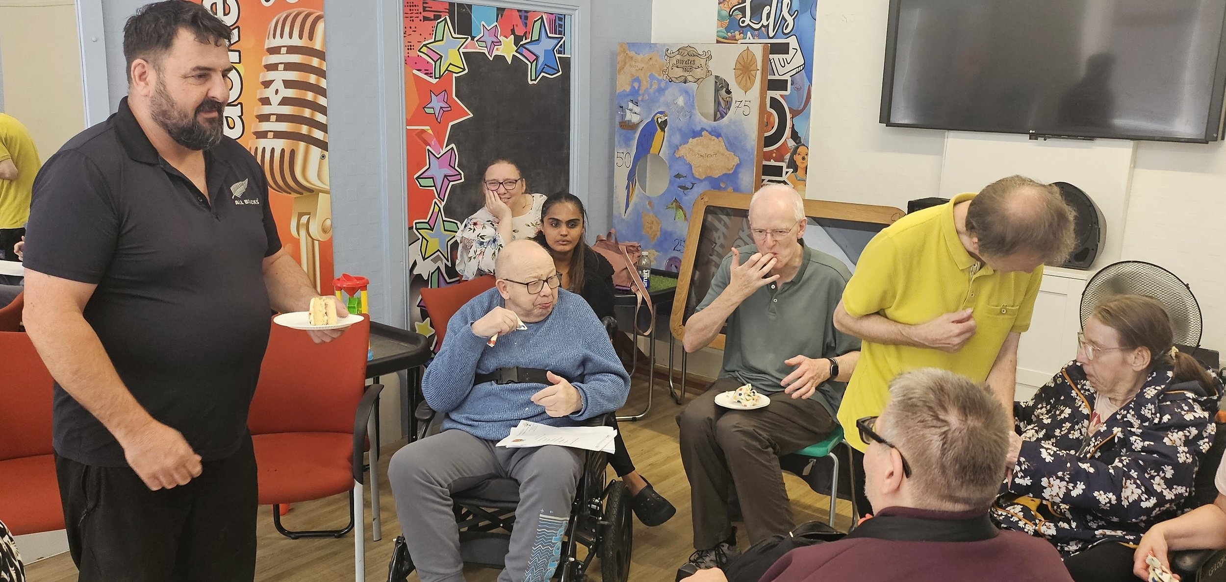A group of people gathered in a room, some sitting and some standing. An older woman in a wheelchair eats cake, a man with glasses and a gray shirt holds his hand to his mouth, and a man in a yellow shirt bends down to talk to a woman with glasses. A