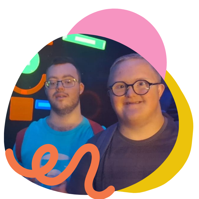 Two men with glasses, one with a beard and blue shirt, the other clean-shaven with a dark shirt, standing together in front of a colorful background at an indoor event.