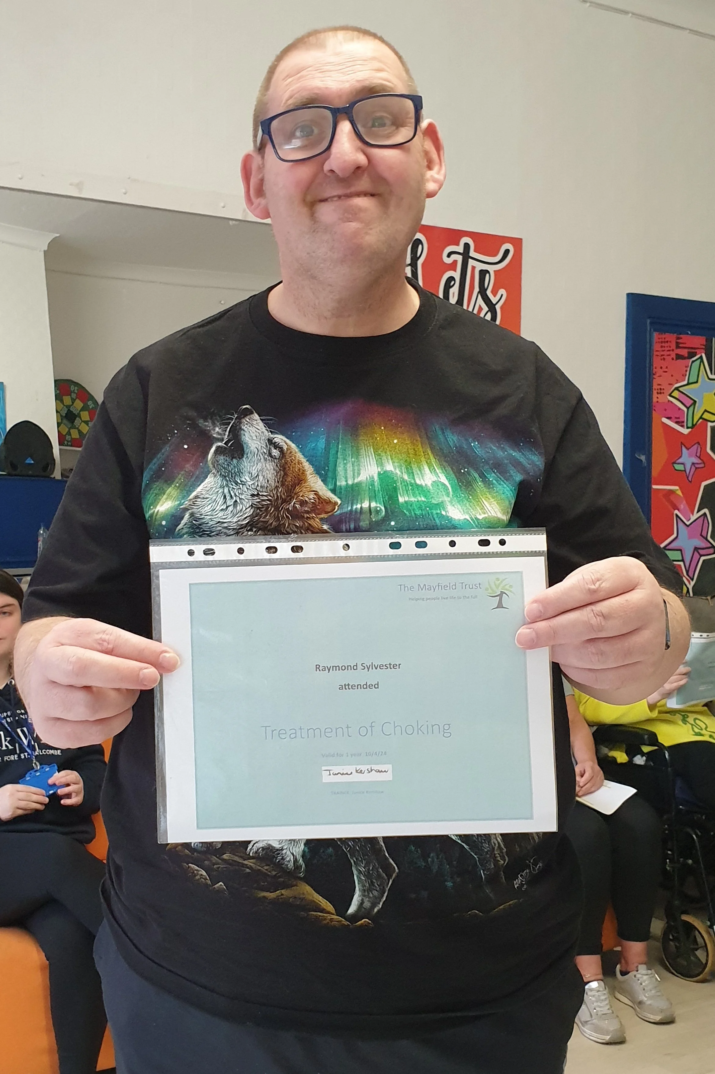 Man holding a certificate for treatment of choking, wearing a black T-shirt with a wolf and colourful light design, in a room with colourful posters and other people in the background.