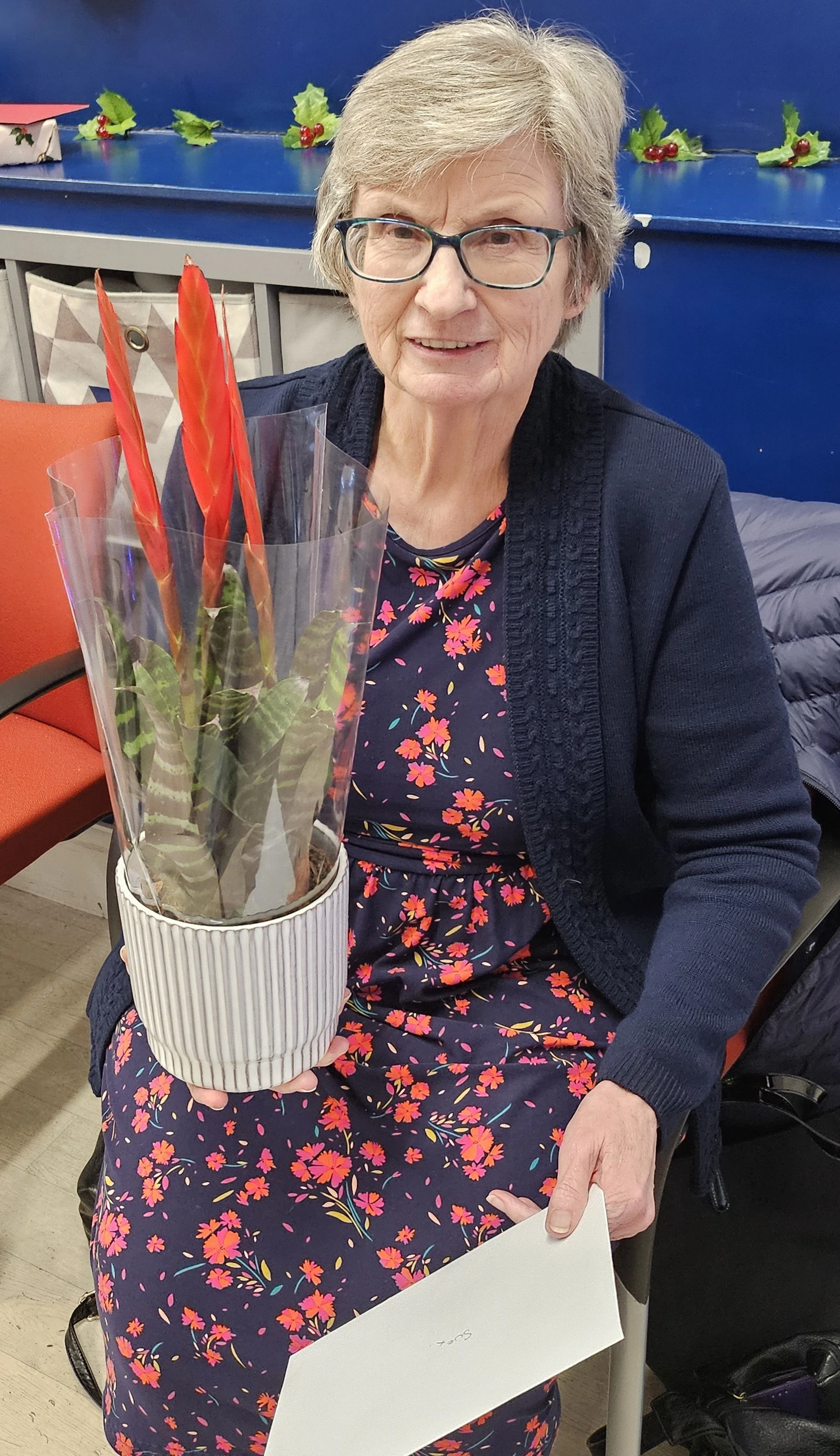A woman with short grey hair and glasses is sitting indoors, holding a potted plant with red exotic flowers. She is wearing a floral dress and a dark cardigan.