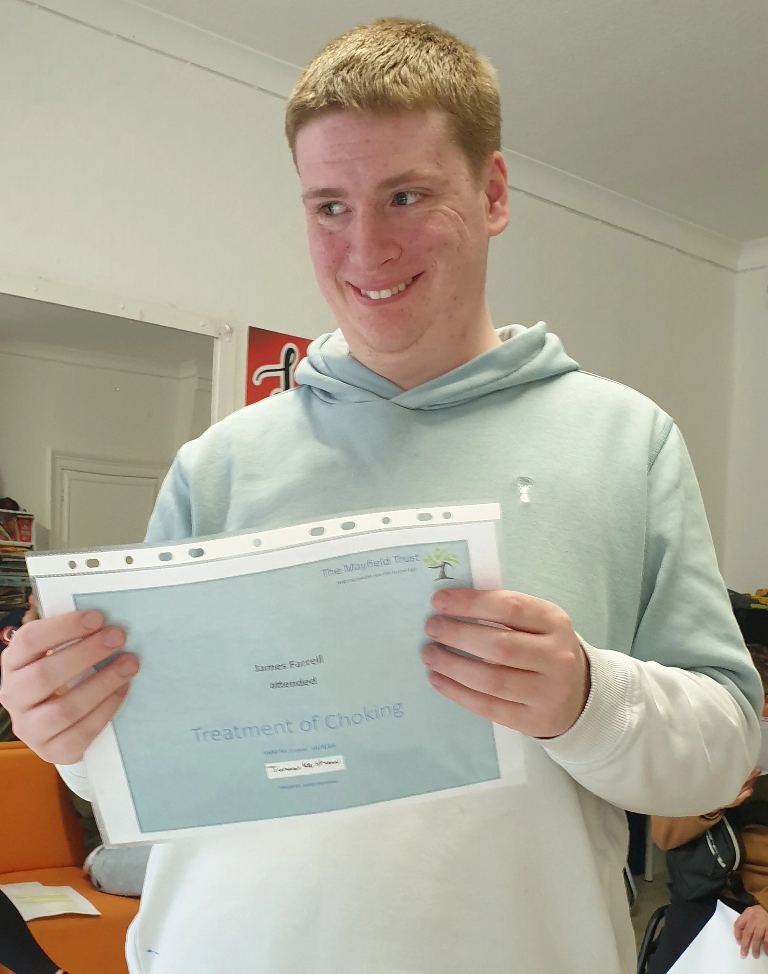 A young man with light brown hair smiling and holding a paper titled 'The Mayfield Trust' at an indoor event. He is wearing a light grey hoodie and has a wedding ring on his left hand. In the background, there are other people and some orange and whi