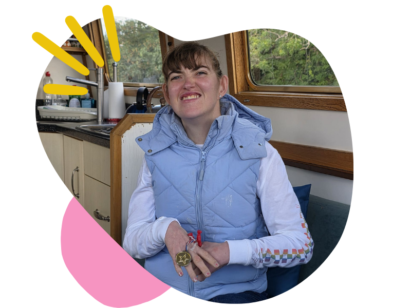 A young woman with brown hair in a ponytail making a grimace inside a camper van, wearing a light blue quilted vest over a white long-sleeve shirt with colorful patterns on the sleeves, and a wristwatch with a star on her wrist.