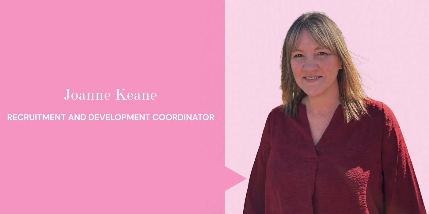 A woman with shoulder-length hair wearing a maroon top, standing outdoors with a pink background.