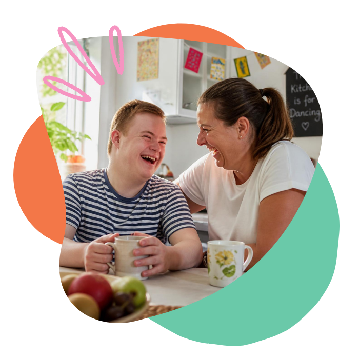 A smiling woman and a boy laughing at a kitchen table holding coffee mugs, with fruits on the table in the foreground.