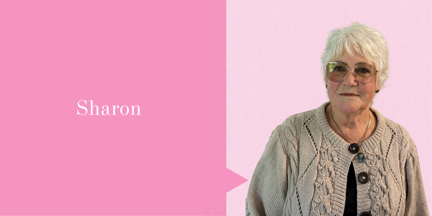 Portrait of an elderly woman with short white hair and glasses, wearing a knitted beige cardigan with large buttons and a necklace, standing against a pink background.