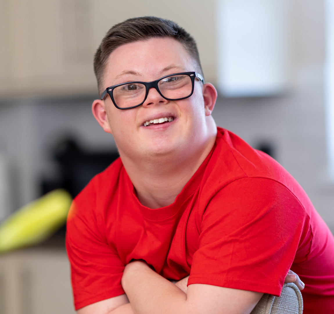 A smiling young man with short brown hair, wearing glasses and a red t-shirt, sitting in a bright room.