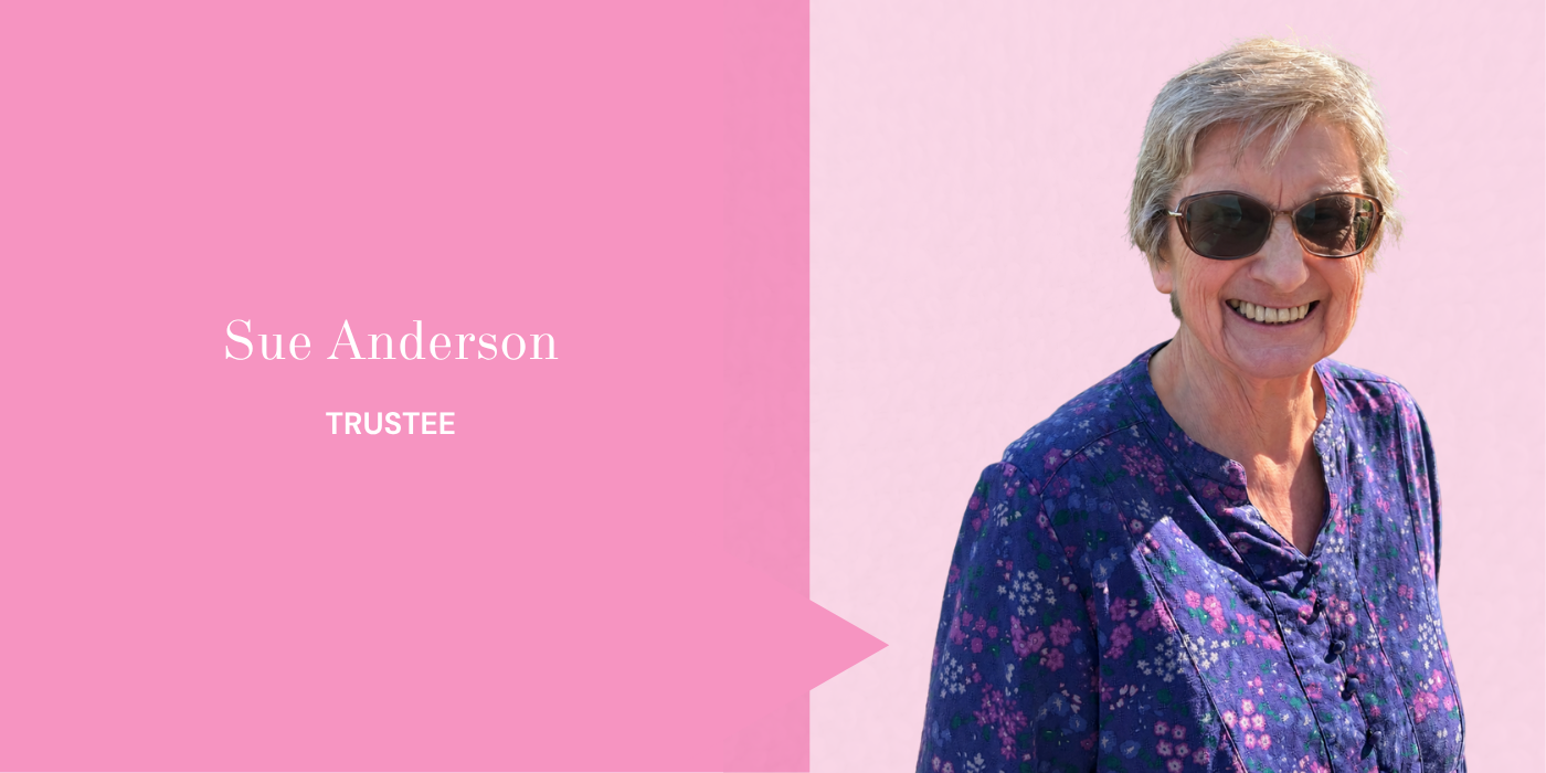 Smiling elderly woman with short gray hair wearing sunglasses and a purple floral blouse, standing in front of a light pink background.