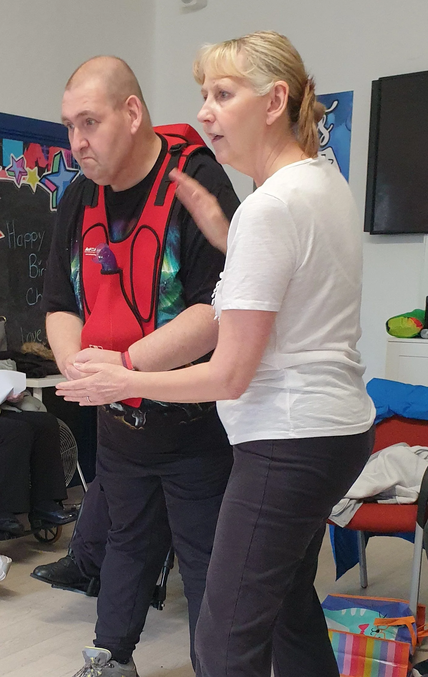 A woman in a white shirt and gray pants is speaking, with her hand near her shoulder, next to a man in a black shirt, black pants, and a red harness, at a birthday celebration with colorful decorations and gifts in the background.