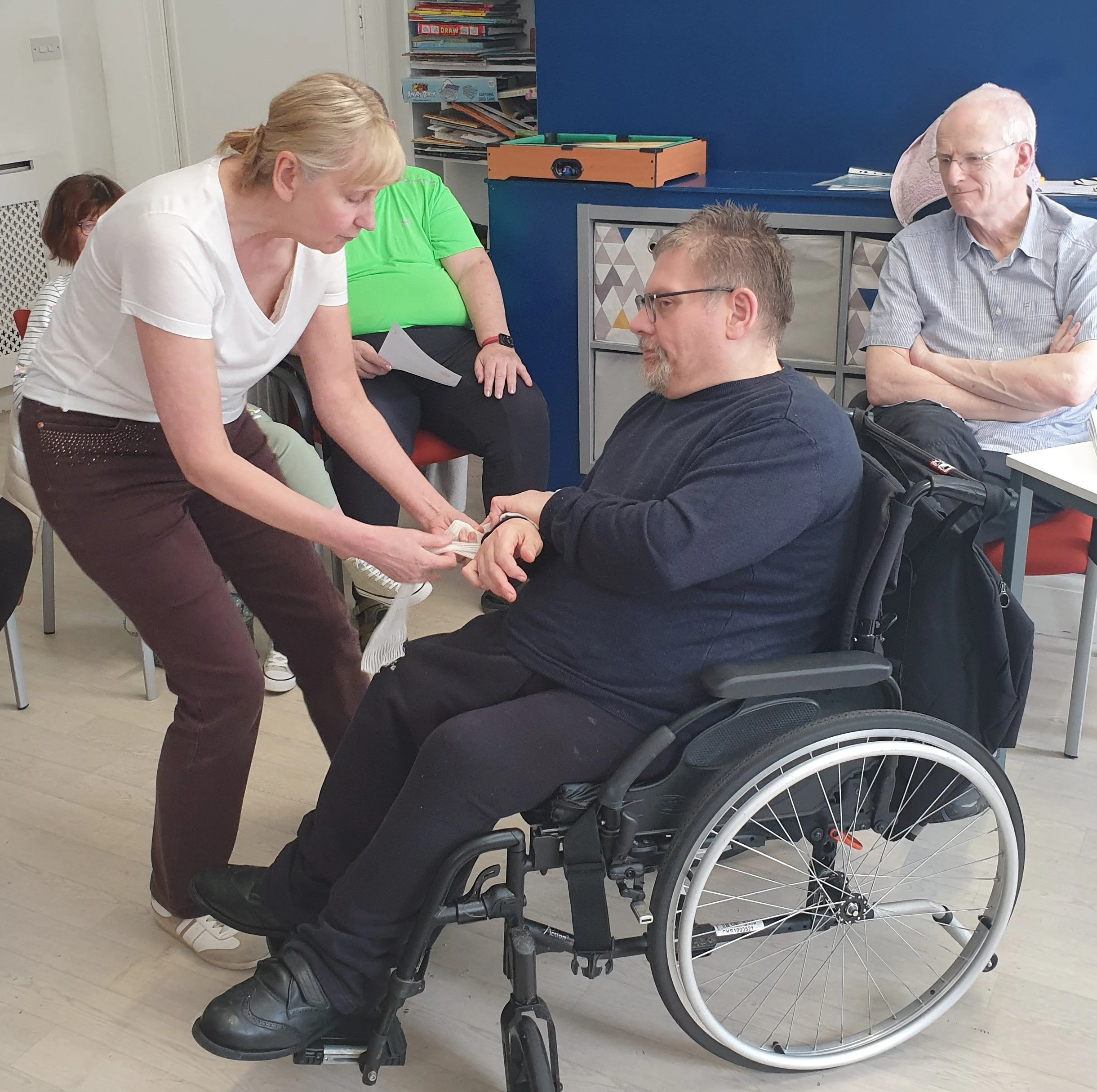 A woman in a white shirt and brown pants is leaning over a man in a wheelchair, holding his hands. The man has gray hair and a beard, and is wearing glasses and a black shirt. In the background, there are people sitting and a bookshelf with books and