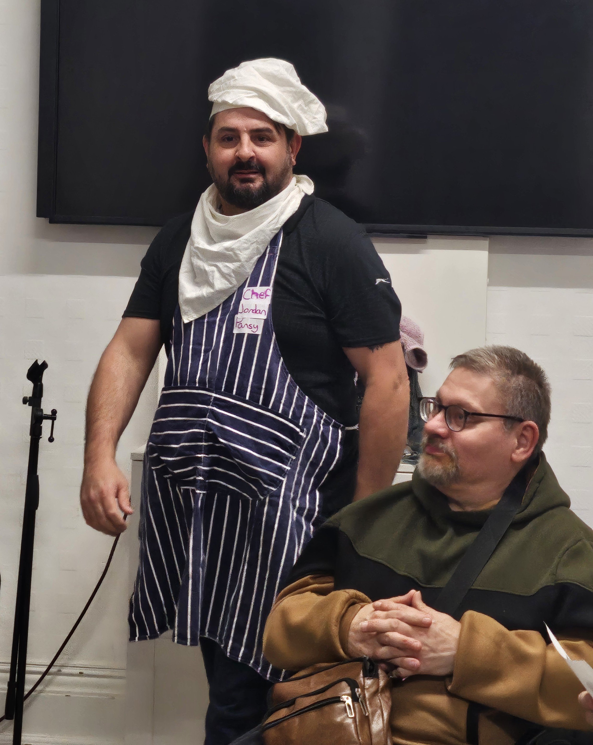 A man with a beard wearing a chef's hat and apron, and a white scarf around his neck, standing indoors against a black wall, next to a seated man with glasses and a beard wearing a brown jacket, in a casual setting.