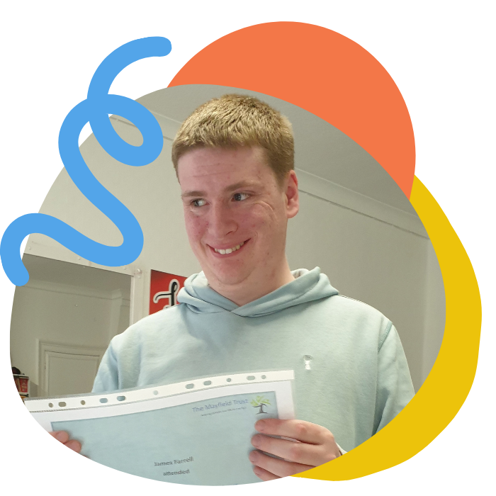 A young man with short red hair smiling and holding a piece of paper in an indoor setting.