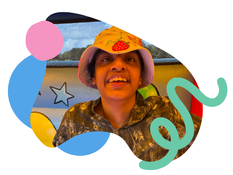 Young man wearing a pink bucket hat with a strawberry design, smiling in front of colorful background with stars and abstract shapes.