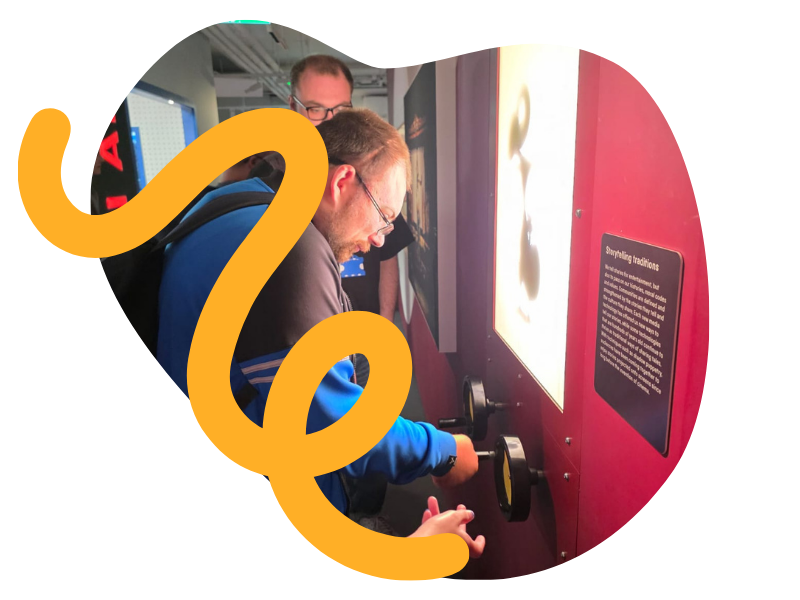 Two young men looking at an interactive exhibit with bright lighting and a red background, one of them operating a knob or dial while the other observes.