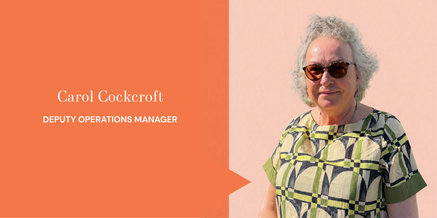 A woman with short curly gray hair wearing sunglasses and a patterned shirt, standing against a light pink background.