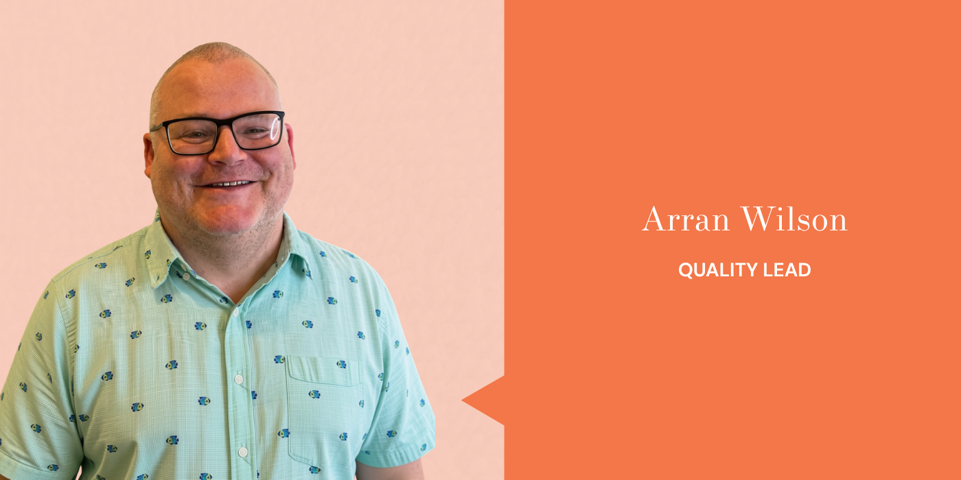 A man with glasses smiling, wearing a light green shirt with small blue and purple patterns, standing against a pink wall. On the right side of the image, an orange speech bubble contains the text: "Arran Wilson, QUALITY LEAD."