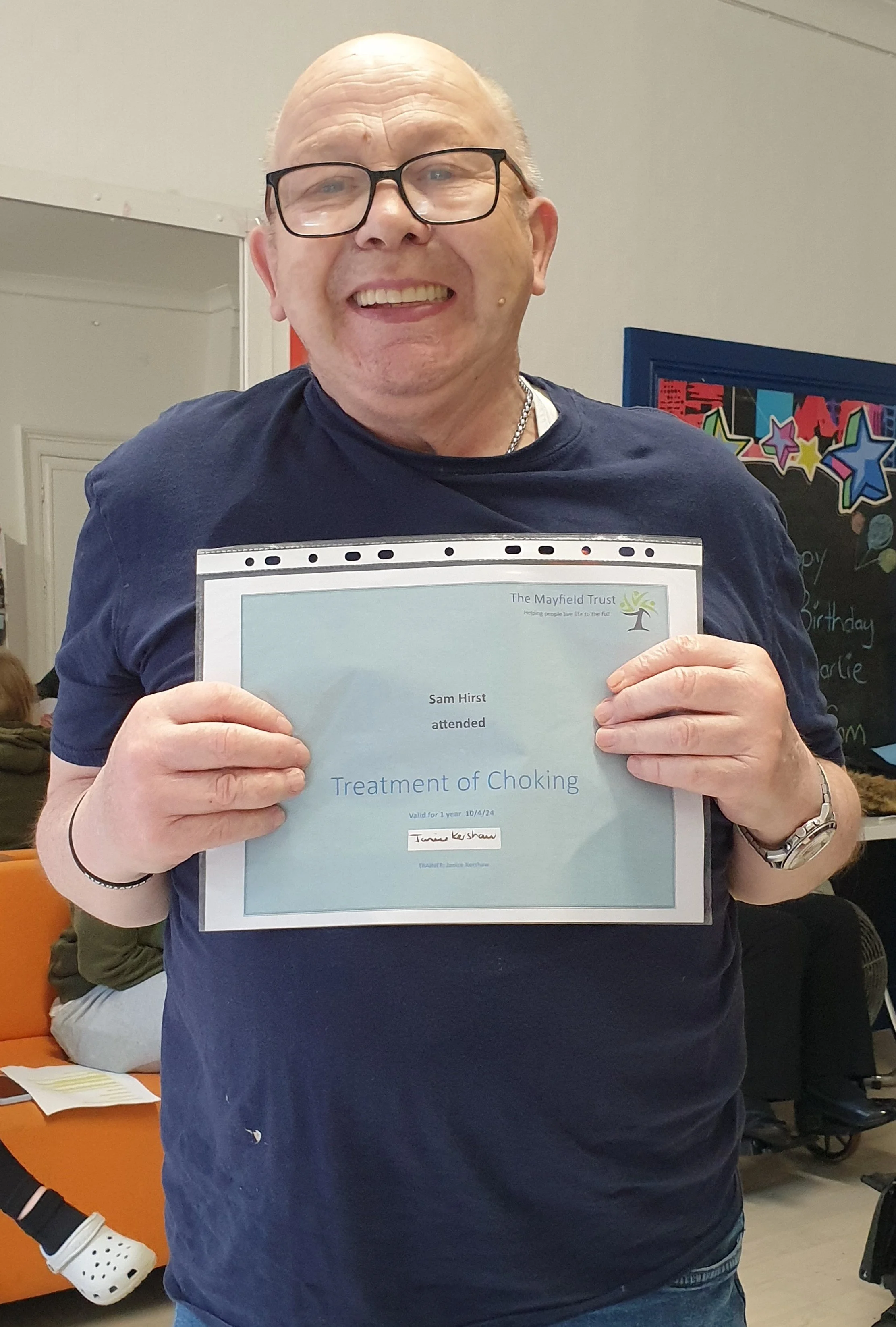 A smiling man with glasses and a chain necklace holding a certificate for treatment of choking, standing in a room with colourful decorations in the background.