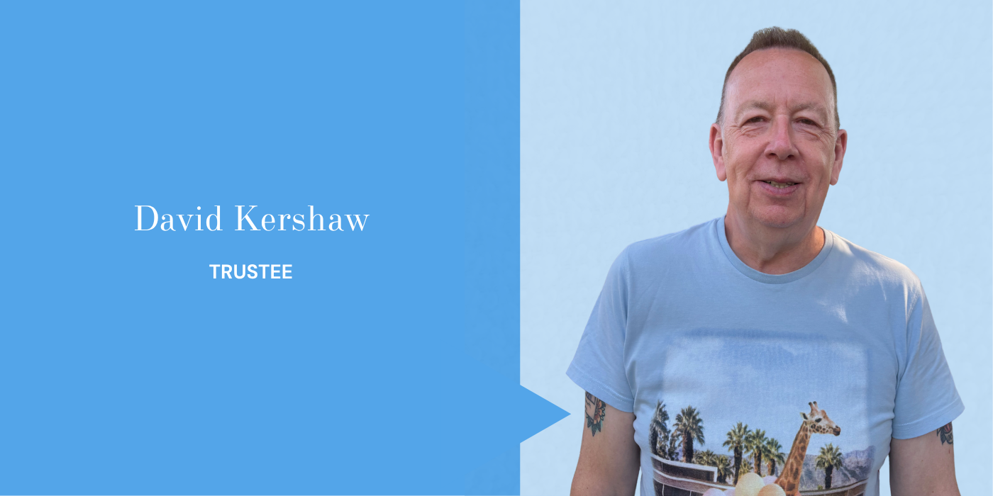 A smiling man wearing a light blue t-shirt with a tropical scene, including palm trees and a giraffe, standing against a light blue background.