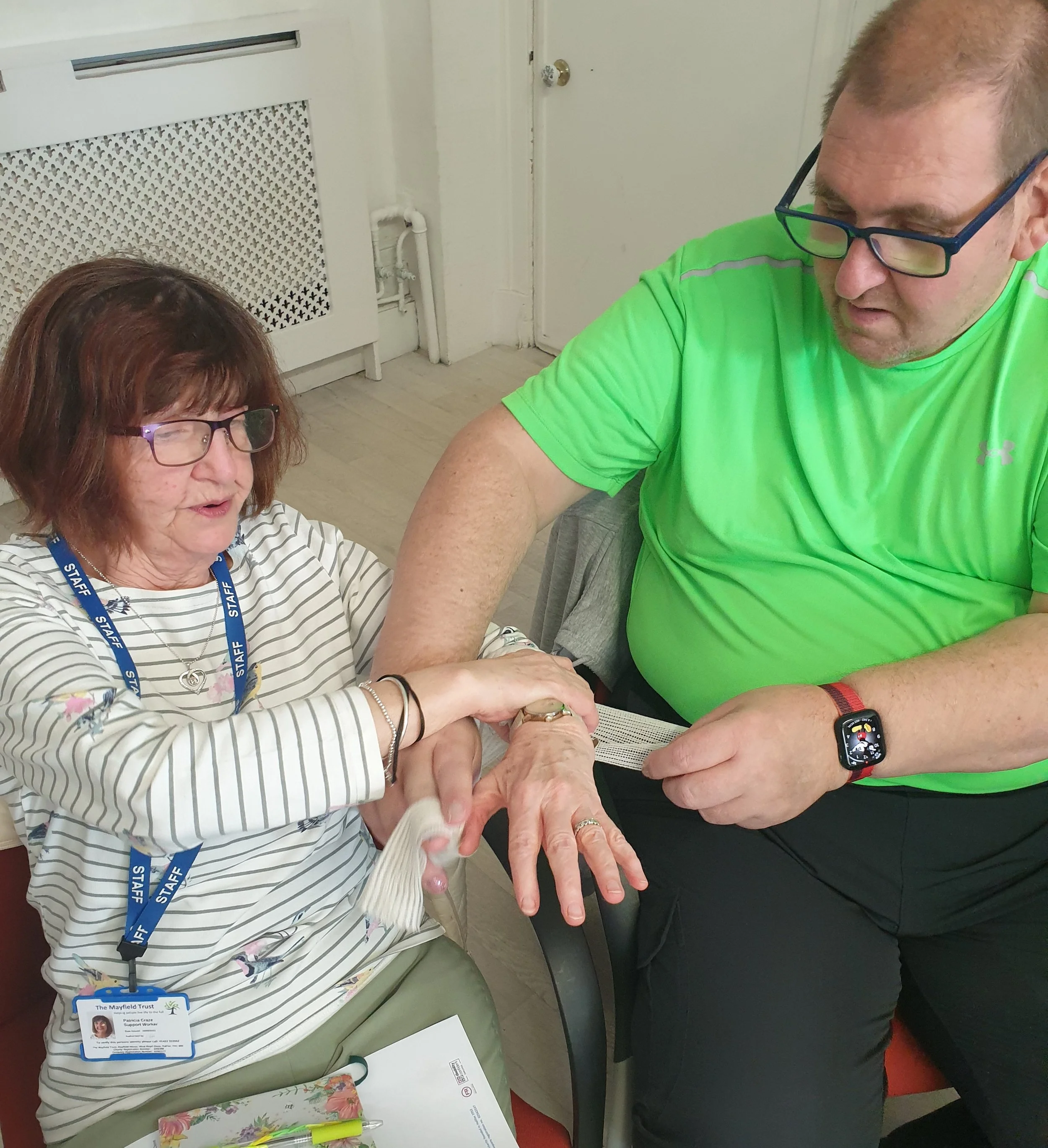 A woman with glasses and a supporter badge with a man wearing glasses and a bright green shirt. They are indoors at a table with documents and writing supplies.