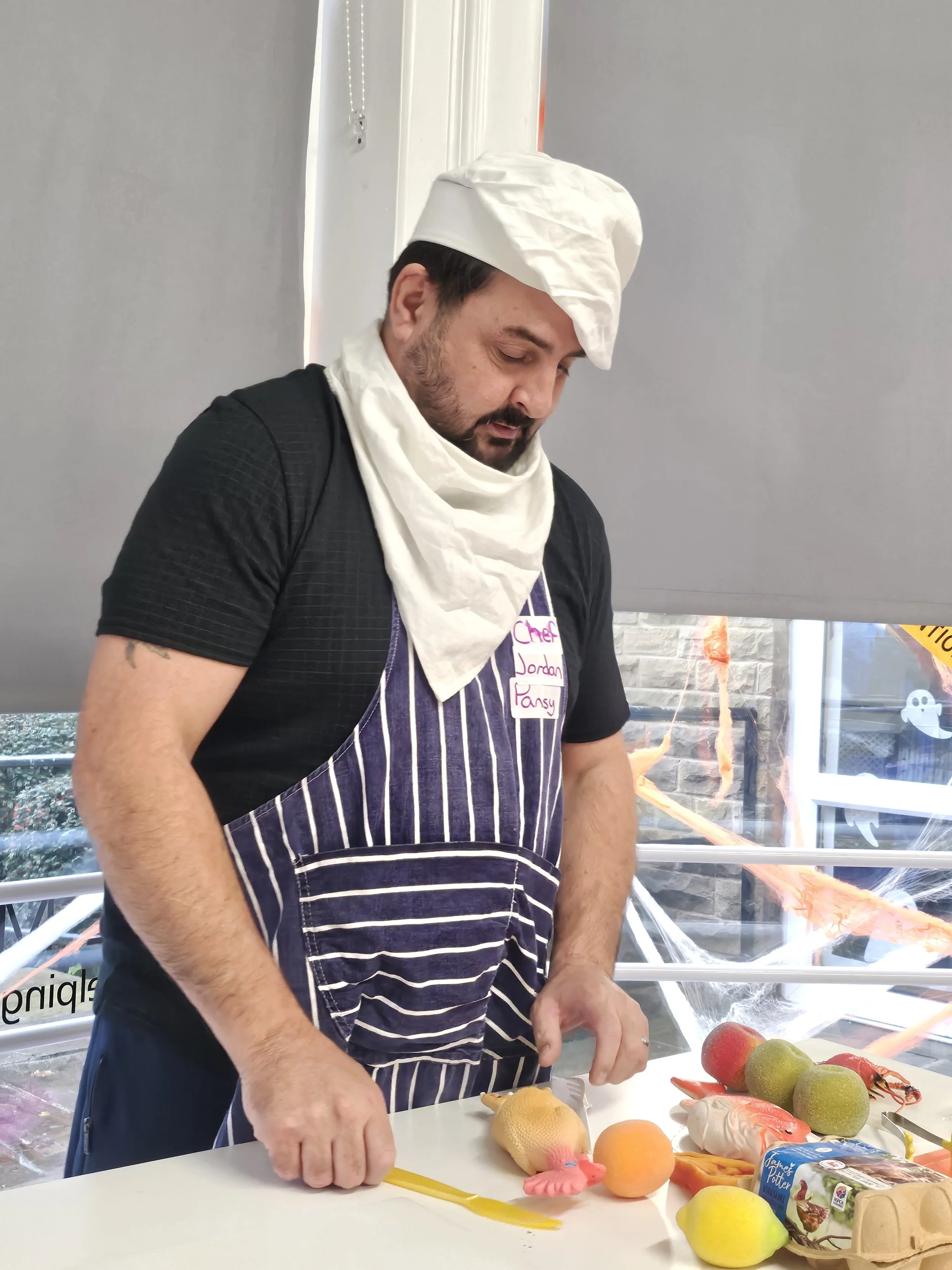 A man wearing a white chef hat and apron with a name tag is preparing food at a table with fruits and fish models, in a brightly lit indoor space with a gray wall and a window showing outdoor scenery.