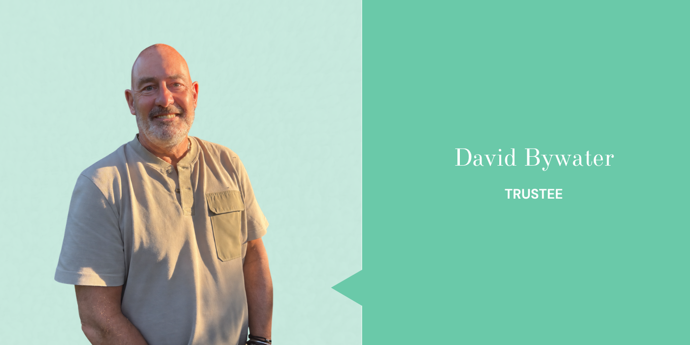 Portrait of a smiling middle-aged man with a beard, wearing a beige shirt, standing against a plain light background, with a green graphic overlay showing his name, David Bywater, and his title, Trustee.