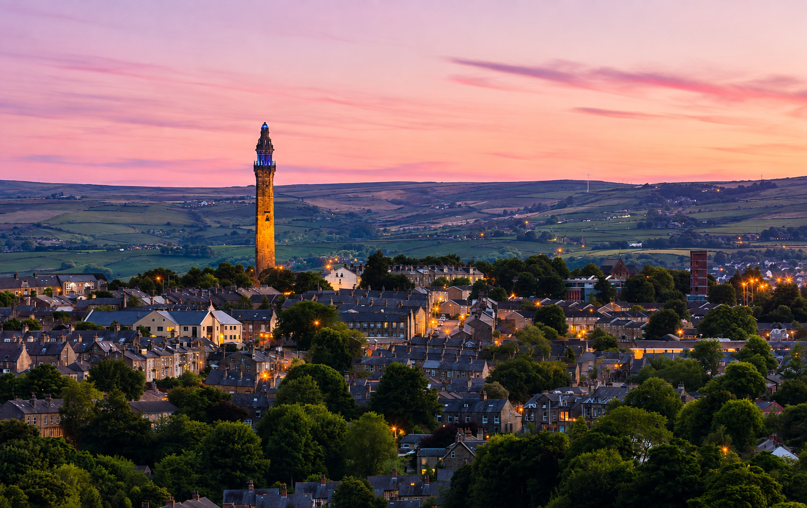 A scenic view of a small town during sunset with a tall historic clock tower in the center, surrounded by houses and greenery, and rolling hills in the background under a colorful sky.