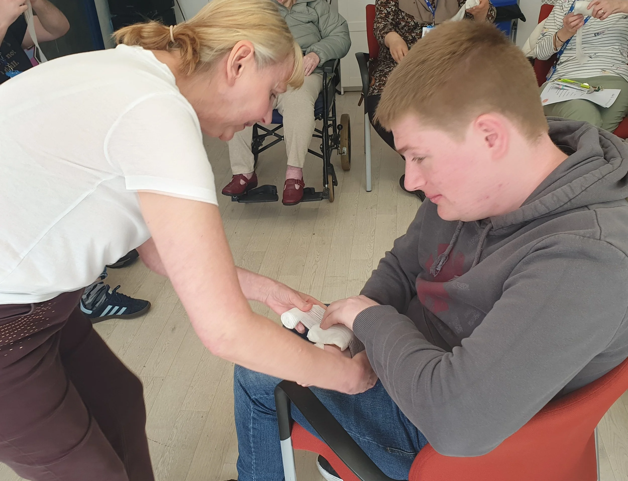 First aid training involves performing bandage wrapping around a hand with several onlookers seated nearby.