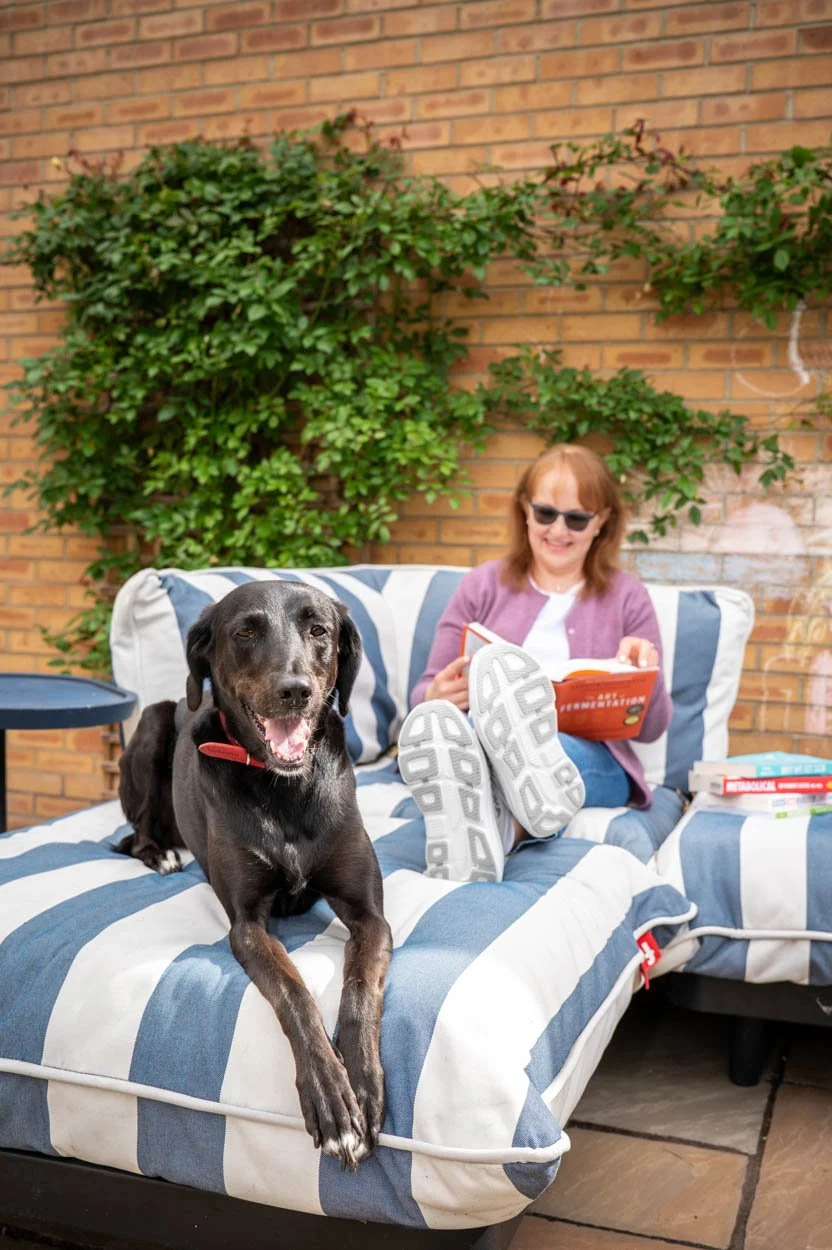 A woman with red hair and sunglasses relaxing on a striped outdoor couch, reading a book, with a black dog lying on the couch in front of her, outdoors against a brick wall and greenery.
