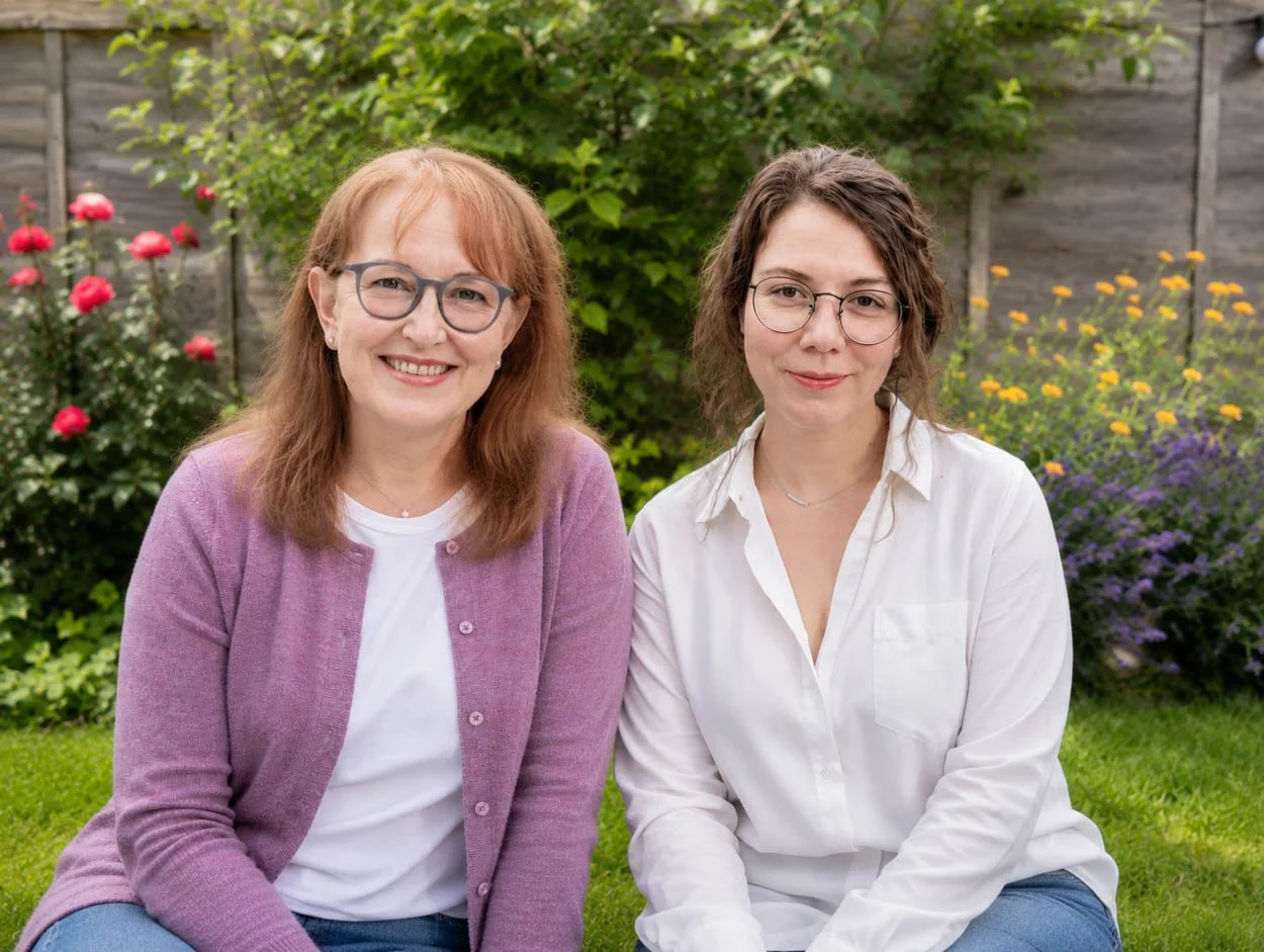 Two women sitting on the grass in a garden with colorful flowers and greenery in the background.